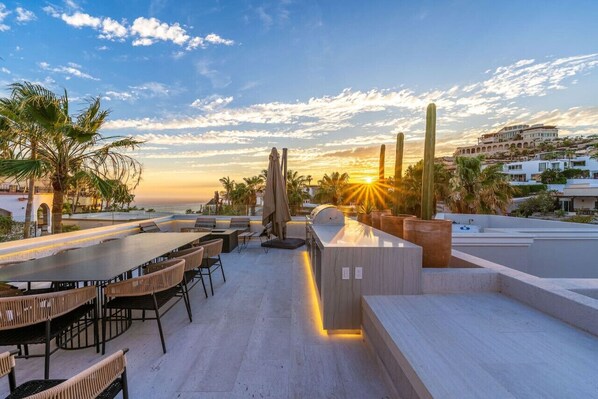 An outdoor dining area with a view of the sunset, palm trees, and a building in the background.