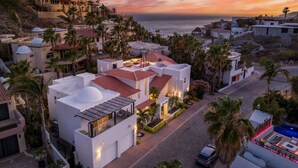 A coastal residential area with modern architecture, including a house with a red-tiled roof and a white facade, surrounded by palm trees and a view of the sea.