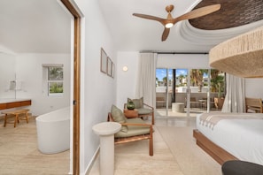 A modern hotel room with a large bed, a sitting area with a chair and a small table, a bathtub, and a ceiling fan. - Casa La Mexicana @ Pedregal Cabos (Cabo San Lucas)
