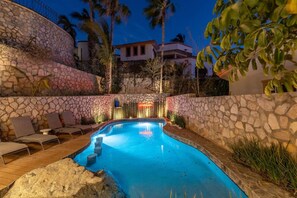 A swimming pool with stone walls and lounge chairs, surrounded by palm trees and a building in the background. - Casa La Mexicana @ Pedregal Cabos (Cabo San Lucas)