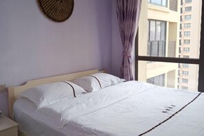 Ein ordentlich bezogenes Bett mit weißen Leinen und einem dekorativen Kopfteil, ein Nachttisch mit Lampe und Blick auf ein Gebäude durch ein Fenster mit violetten Vorhängen.