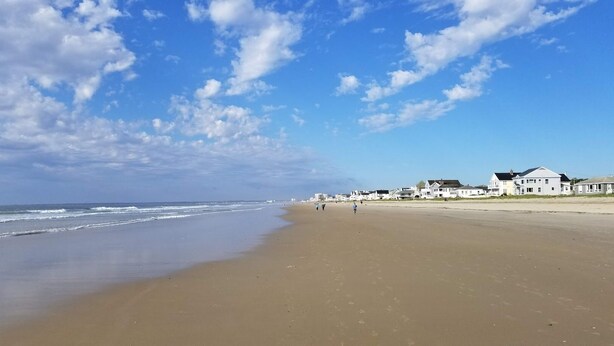 A sandy beach with a few people walking, a clear sky, and houses in the background.