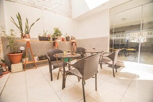 Un patio soleado con sillas y mesas de mimbre, rodeado de plantas en macetas.