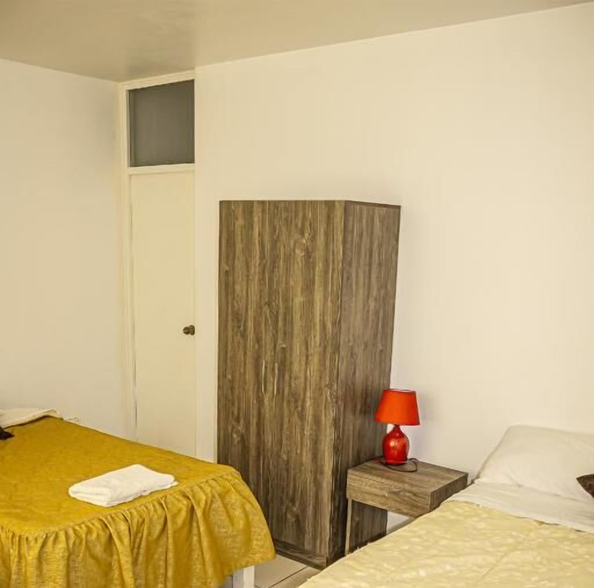 A room with two beds, one with a yellow bedspread and the other with a white bedspread. There is a wooden wardrobe and a red lamp on a wooden nightstand.