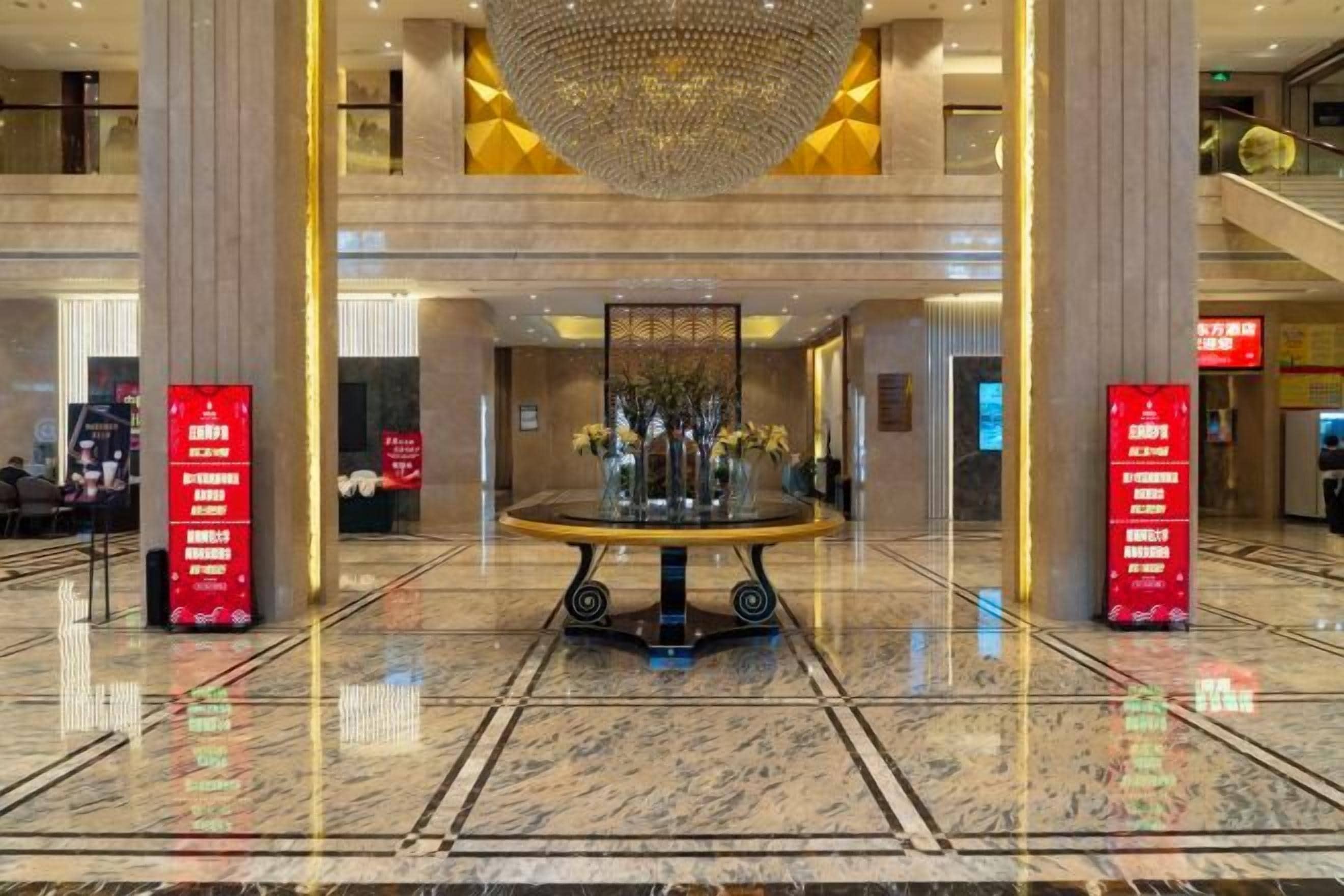 A hotel lobby with a central round table, two red vertical signs, and a large chandelier.
