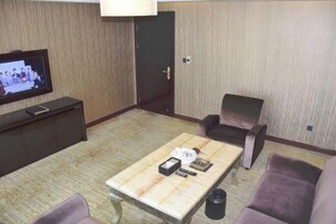 Habitación de hotel con televisión de pantalla plana sobre un soporte, una mesa de centro con un control remoto, una revista y una caja de pañuelos. Hay dos sillones y un sofá.