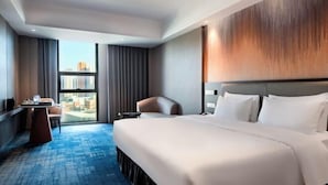 Habitación de hotel con una cama grande, un escritorio, una silla y vista a la ciudad a través de un ventanal amplio.