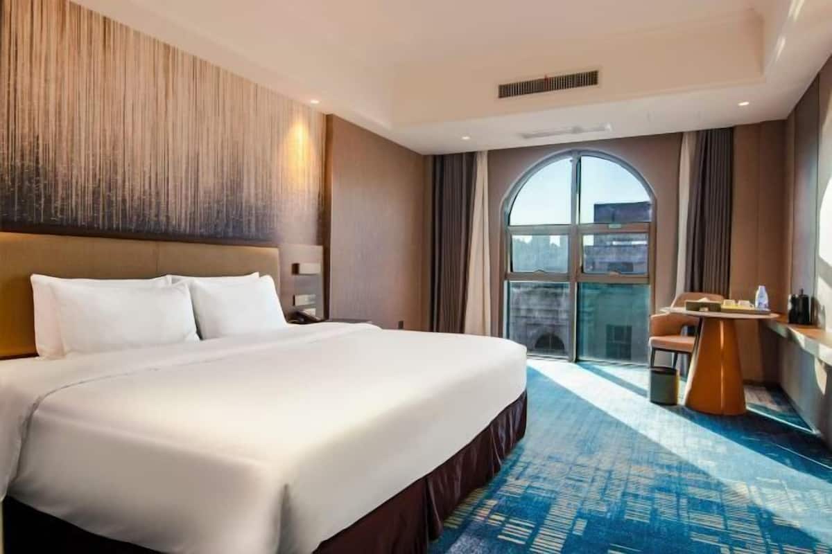 A hotel room with a large bed, a small table, and a view of the city through a large window.
