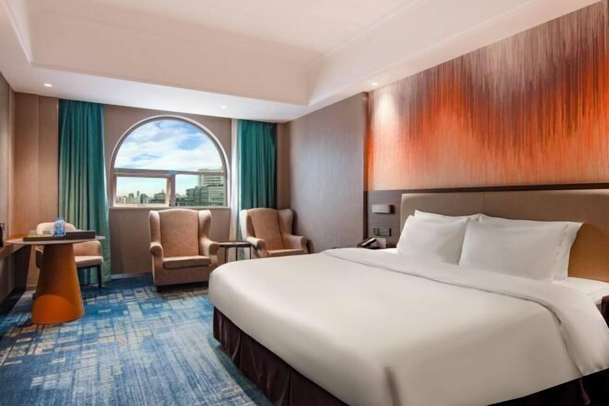 A hotel room with a large bed, two armchairs, a small table, and a view of the city through a window.
