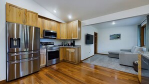 A modern kitchen with stainless steel appliances, wooden cabinets, and a grey carpeted living area with a grey sofa and a flat-screen TV.