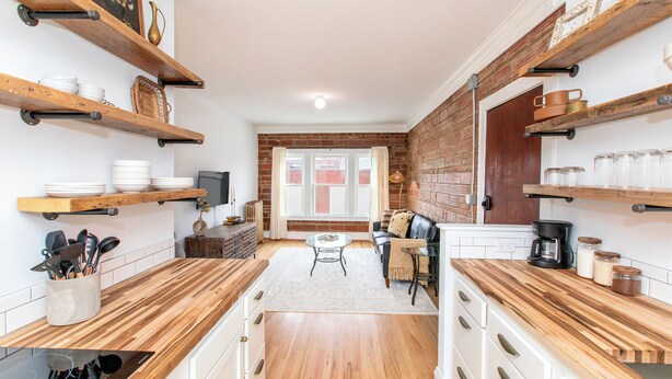 A kitchen with wooden countertops, white cabinets, and brick walls. There are shelves with dishes and jars, a coffee maker, and a dining area with a sofa and table. - Modern Loft in Historic Birdtown - Unit 6 (Lakewood)