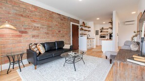 A modern living room with a brick wall, a black leather sofa, a glass coffee table, and a wooden side table. - Modern Loft in Historic Birdtown - Unit 6 (Lakewood)