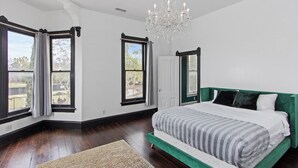 A bedroom with a large bed, dark wooden flooring, a chandelier, and large windows with curtains.