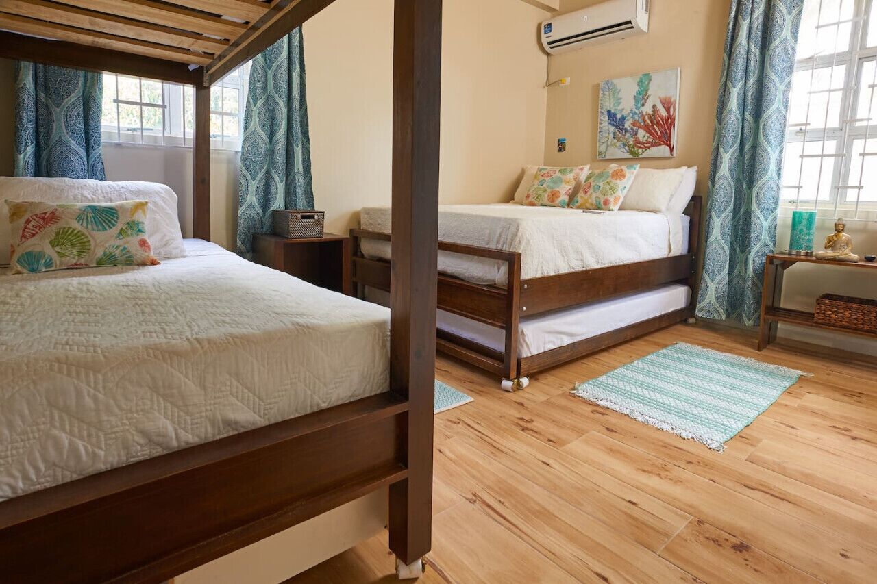 A bedroom with two wooden beds, a window with curtains, an air conditioning unit, and a painting on the wall.