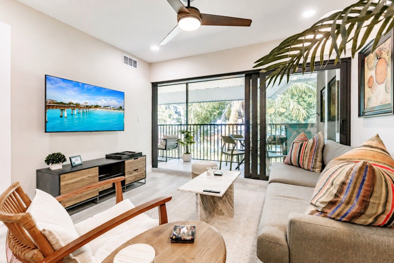 A modern living room with a large TV, comfortable seating, a balcony with outdoor furniture, and a ceiling fan.