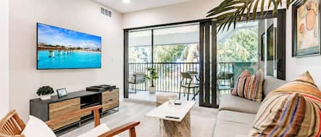 A modern living room with a large TV, comfortable seating, a balcony with outdoor furniture, and a ceiling fan.