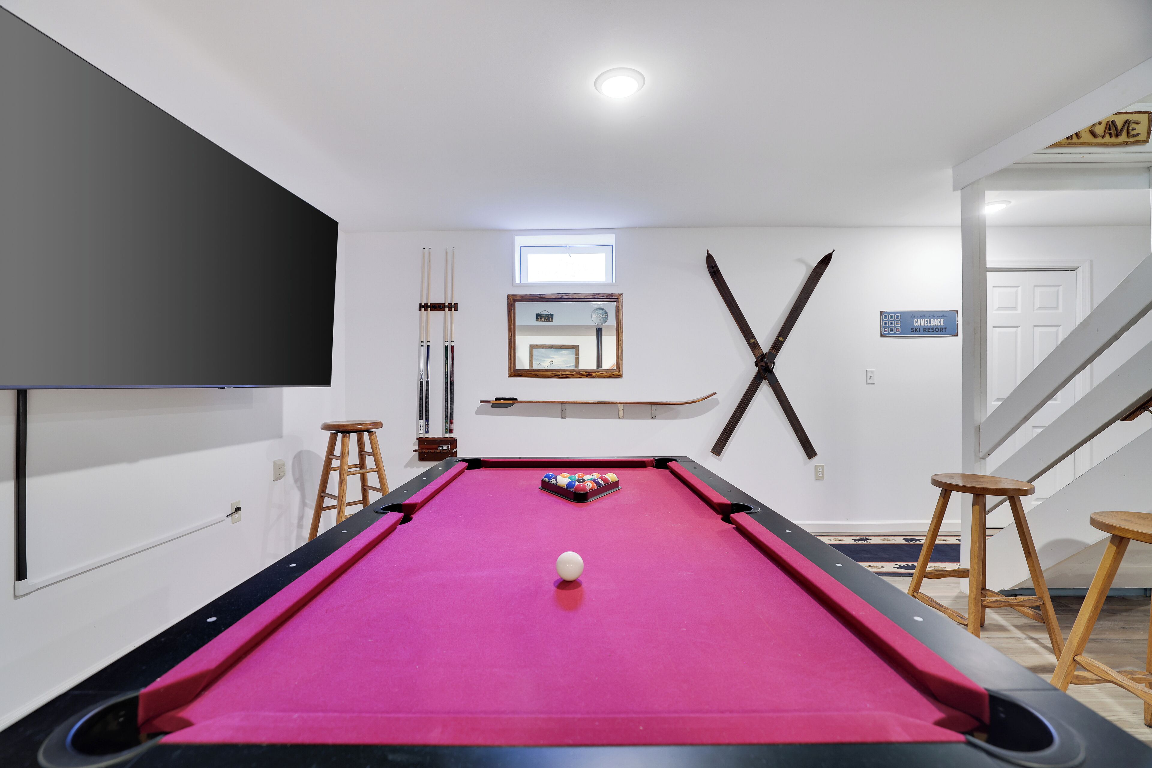 A pool table with a pink felt surface, a white ball in the center, and pool cues on the left. There is a window above the table and a sign on the wall to the right.
