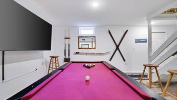 A pool table with a pink felt surface, a white ball in the center, and pool cues on the left. There is a window above the table and a sign on the wall to the right.