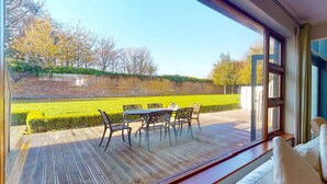 A room with a large window overlooking a neatly maintained garden with a patio table and chairs. - Castlemartyr Holiday Lodges No 3 (2 Bed) (Castlemartyr)
