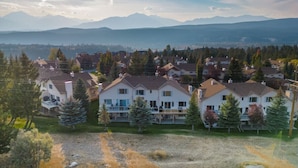A residential area with multi-story houses, some with balconies, surrounded by trees and a mountainous backdrop.