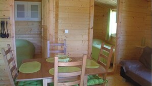 A wooden cabin with a dining table set for four, green cushioned chairs, a wooden sofa, and a cabinet with glass doors.