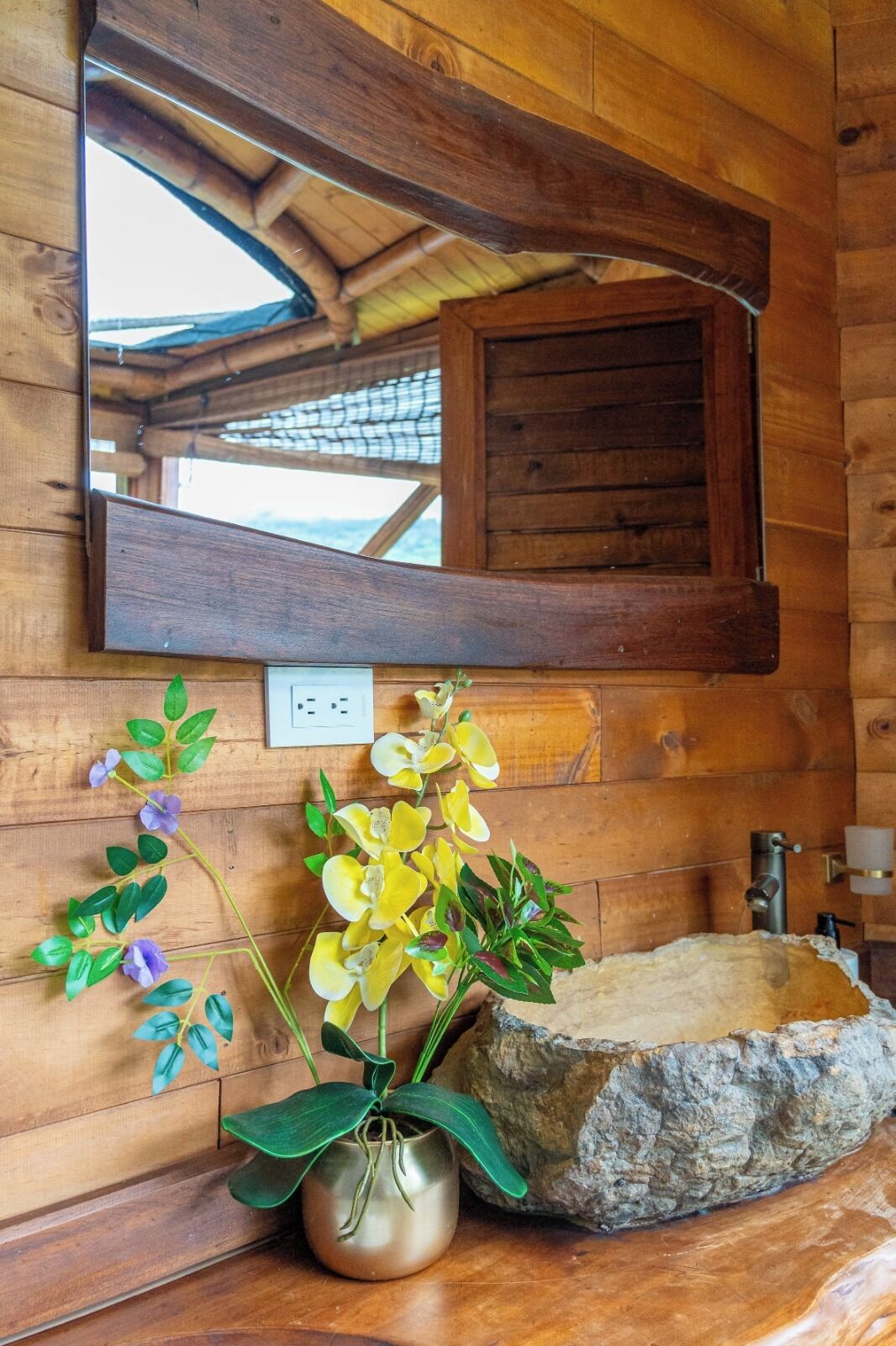 A wooden cabin interior with a stone sink, a mirror above it, and a potted plant with yellow and purple flowers.