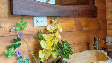 A wooden cabin interior with a stone sink, a mirror above it, and a potted plant with yellow and purple flowers.