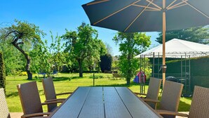 Een terras met een tafel en stoelen, een grote parasol en uitzicht op een gazon met bomen en een witte tent.