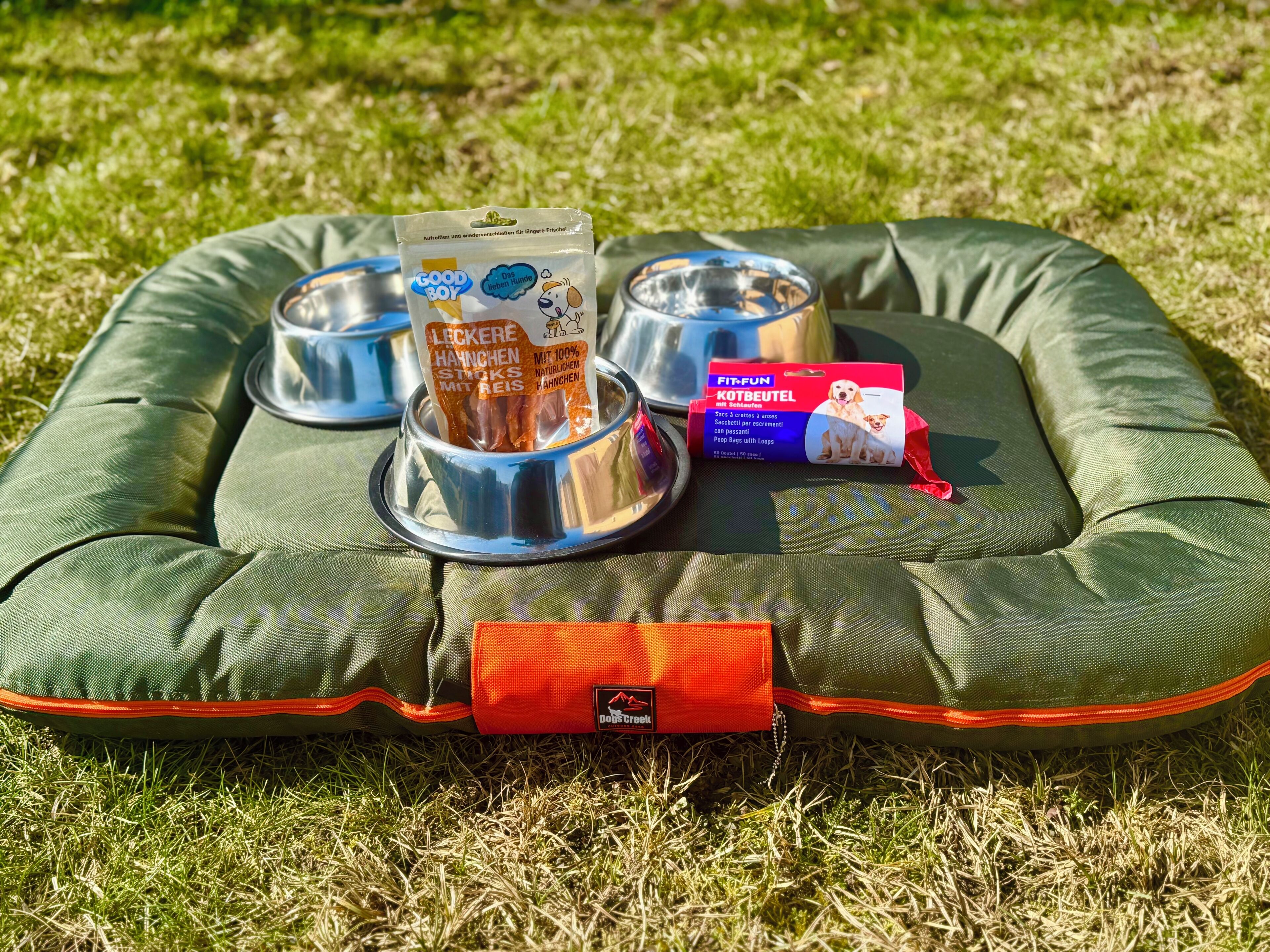 A dog bed with two bowls and a pack of dog food on a grassy area.