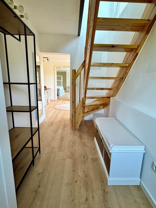 Un couloir étroit avec un escalier en bois menant à un étage supérieur, un banc blanc et une étagère en métal noir.