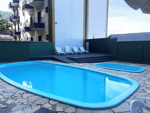 Une piscine divisée en deux zones contiguës, un ensemble de chaises longues blanches et, au fond, un bâtiment doté de balcons.