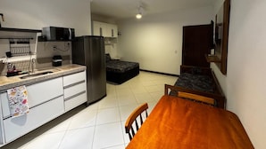 A compact kitchen with a refrigerator, microwave, and sink. There is a dining table with chairs and a small bed in the background. - Dell Mar Rentals - Flats and Apartments for Season - Ubatuba (Ubatuba)
