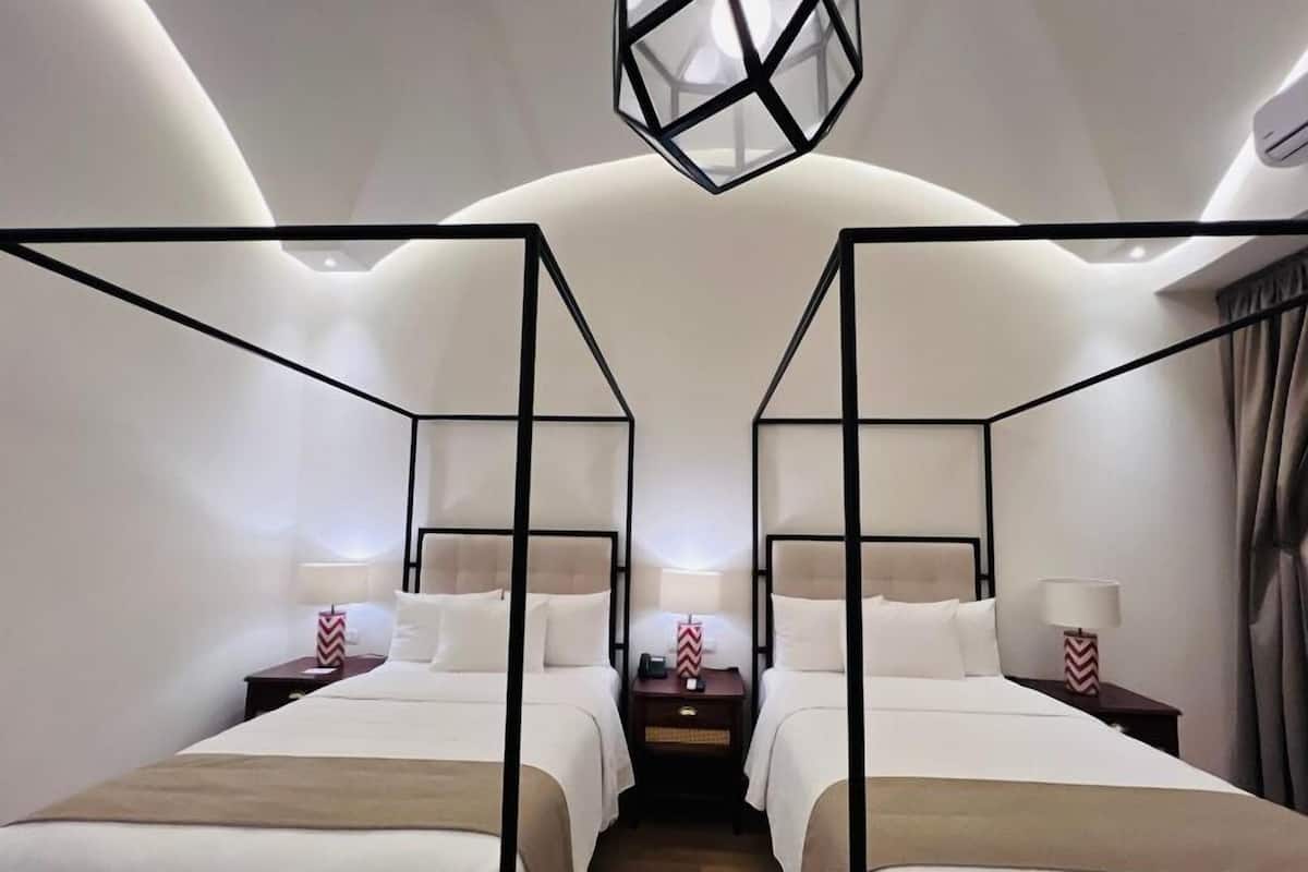 A hotel room with two beds, each with a black metal frame, white bedding, and a small bedside table with a lamp.
