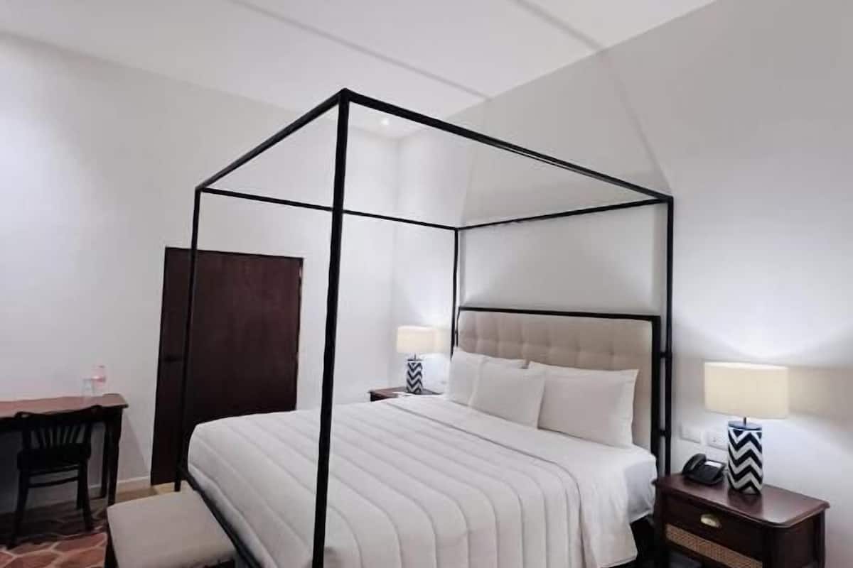 A four-poster bed with a white bedspread and pillows, a wooden nightstand with a lamp, and a chair.