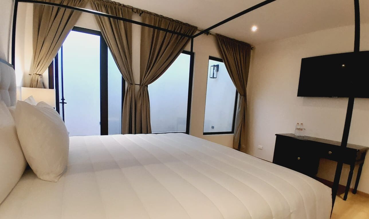 A hotel room with a large bed, a flat-screen TV, a small table, and a view of the sky through the windows.