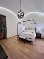 Een moderne slaapkamer met een hemelbed, een houten bankje, een nachtkastje en een geometrische hanglamp.