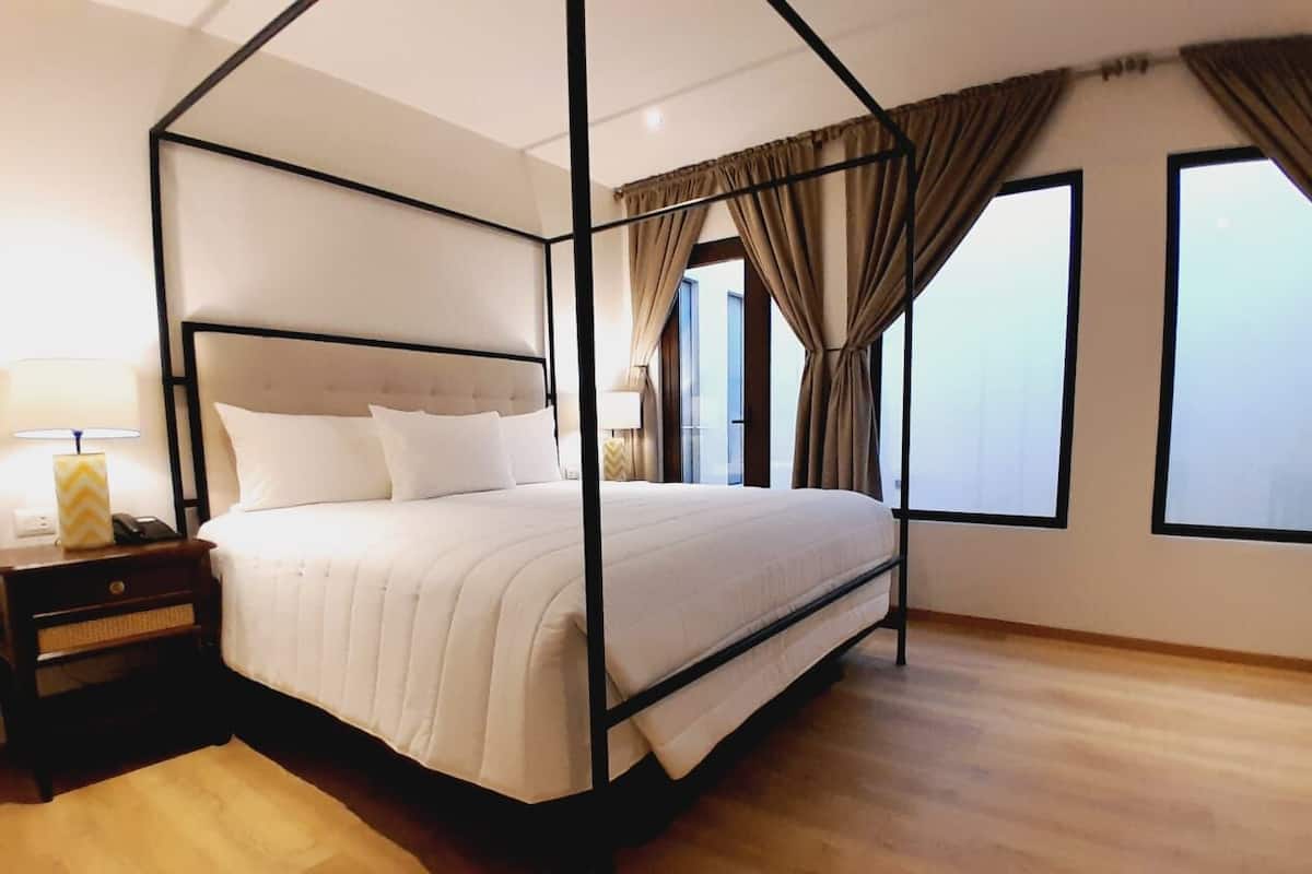A four-poster bed with a white bedspread and pillows, a wooden nightstand with a lamp, and large windows with curtains.