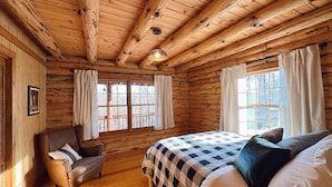 A log cabin bedroom with a bed, a chair, and two windows with curtains. - Charming log cabin by Berkeley Springs w/ hot tub! (Hedgesville)