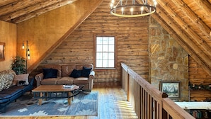 A cozy log cabin interior with a wooden ceiling, a stone fireplace, a leather sofa, and a chandelier. - Charming log cabin by Berkeley Springs w/ hot tub! (Hedgesville)