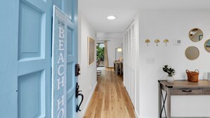 Un couloir avec une porte bleue sur laquelle on peut lire « BIENVENUE À LA PLAGE », un plancher en bois, un bureau avec une plante et un miroir accroché au mur.