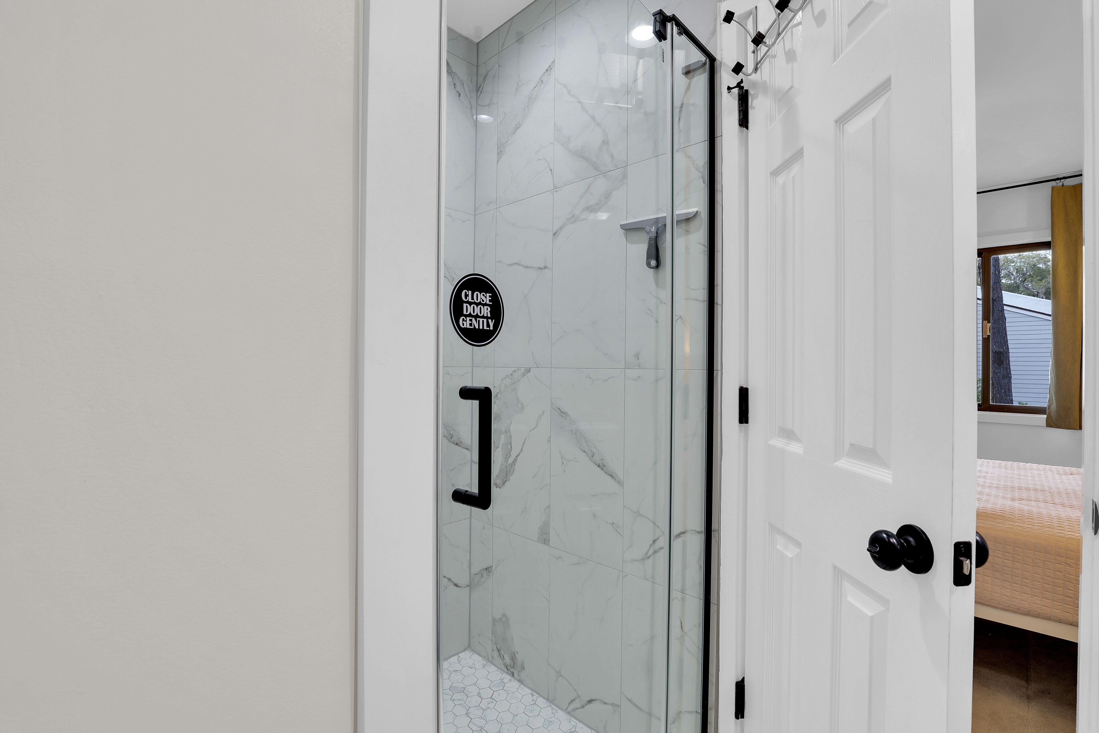 Une salle de bain moderne avec une douche à parois de verre, une pancarte indiquant de « fermer la porte en douceur » et un lit visible dans la pièce adjacente.