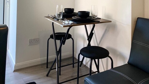 Een eetgedeelte met een tafel gedekt voor twee personen, zwarte stoelen, een zwarte bank, een spiegel en planken met planten.