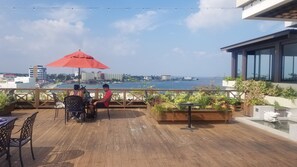 A wooden deck with outdoor seating, a red umbrella, and a view of the sea and cityscape.
