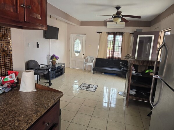 A kitchen with a tiled floor, a ceiling fan, a refrigerator, a television, and a dining area with a sofa and chairs.