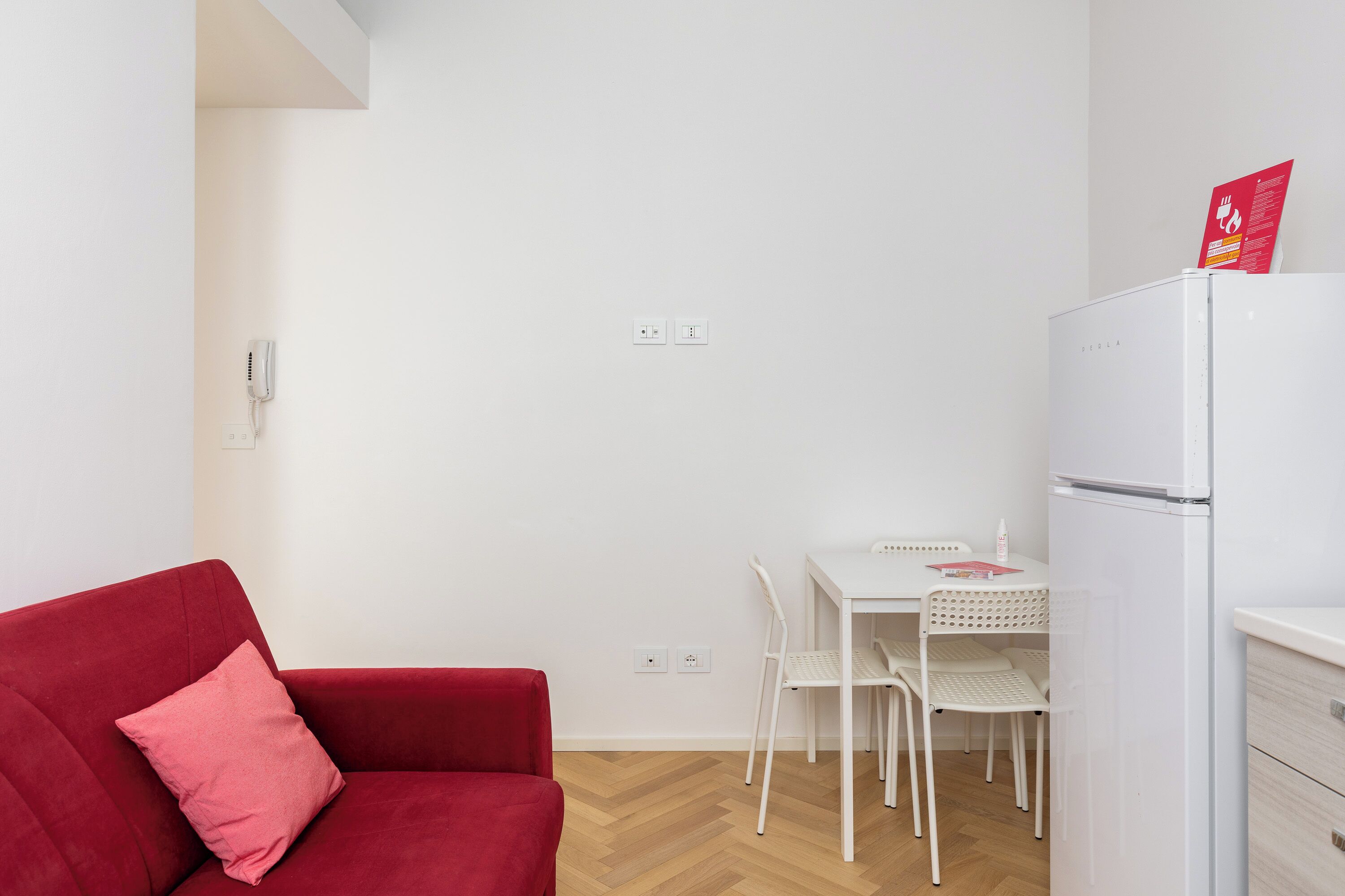 Et lille, moderne værelse med en rød sofa, et hvidt spisebord med stole, et hvidt køleskab og en hvid væg med en stikkontakt.