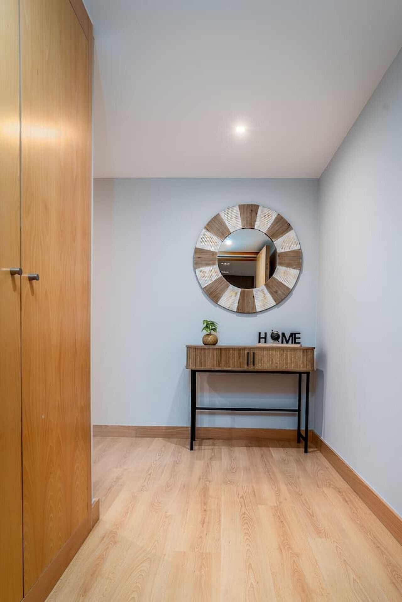 A hallway with a wooden wardrobe, a round mirror with a striped pattern, and a small wooden console table with a plant and a sign that reads 'HOME'.