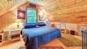 Ein gemütliches Schlafzimmer im Dachgeschoss mit Holzbalkendecke, einem großen Bett mit blauer Bettwäsche, einem Fenster mit Vorhängen und einem farbenfrohen, abstrakten Gemälde.