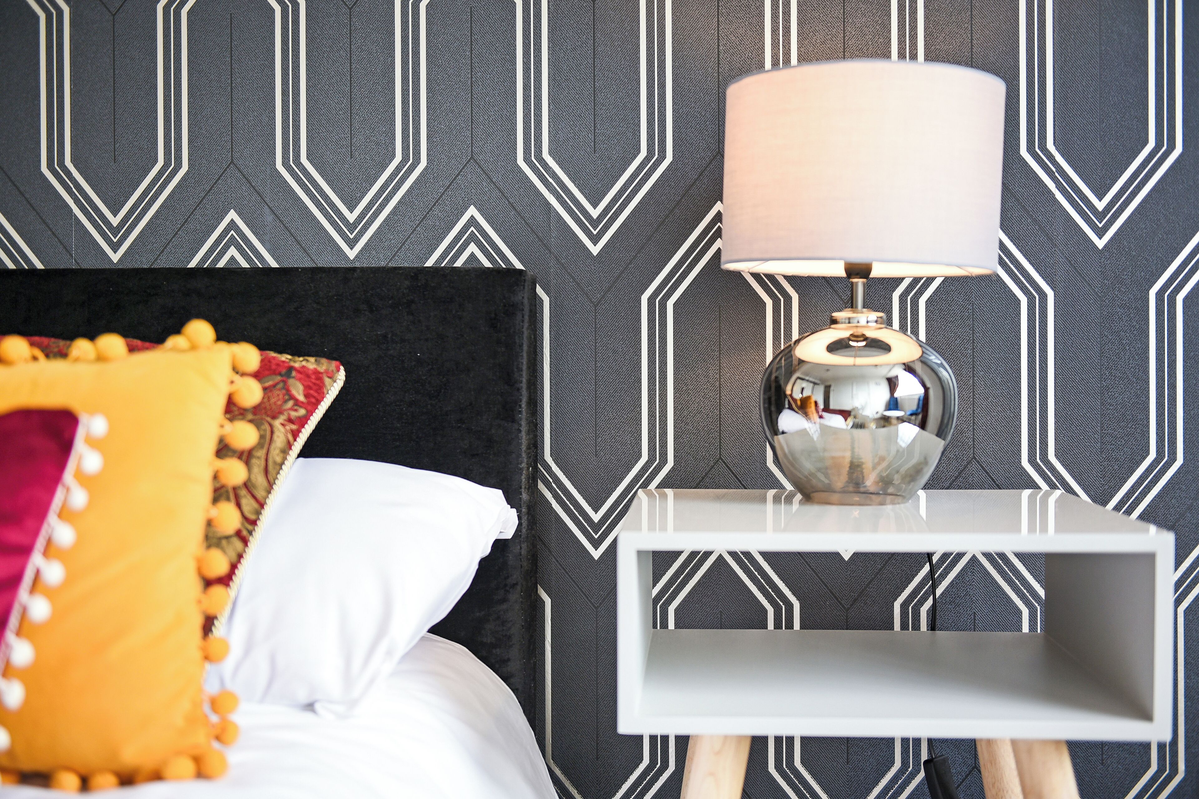 A neatly made bed with a black headboard, a white pillow, and a yellow pillow with pom-poms. There is a white nightstand with a lamp and a small shelf. The wall has a geometric pattern.