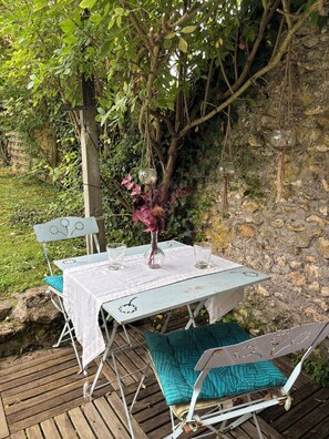 An outdoor dining area with a table set for two, surrounded by greenery and a stone wall. - Maisonnette à la Campagne (Baulne)