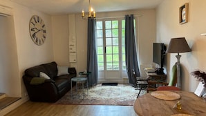 Un salon avec un canapé en cuir marron, une petite table ronde, une table basse en bois, une horloge au mur et une fenêtre avec des rideaux.